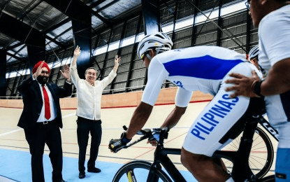 A cyclist wearing a jersey with 'PILIPINAS' on it prepares to ride on a track, while two men, one in a suit and the other in a traditional outfit, cheer and raise their hands in excitement.