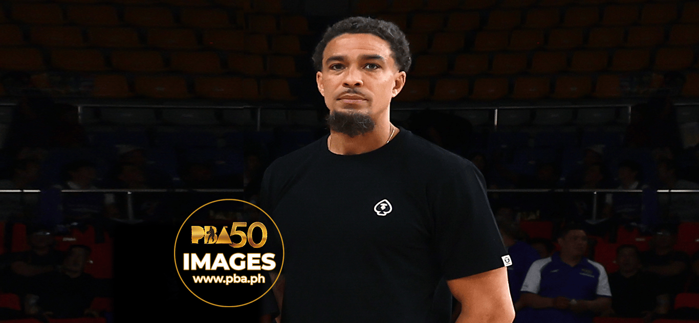 A basketball player wearing a black t-shirt stands on the court, with an audience in the background.