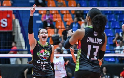 A volleyball player in a black jersey celebrates energetically with raised arms while another player wearing a similar jersey looks on, both on a court with a volleyball net in the background.