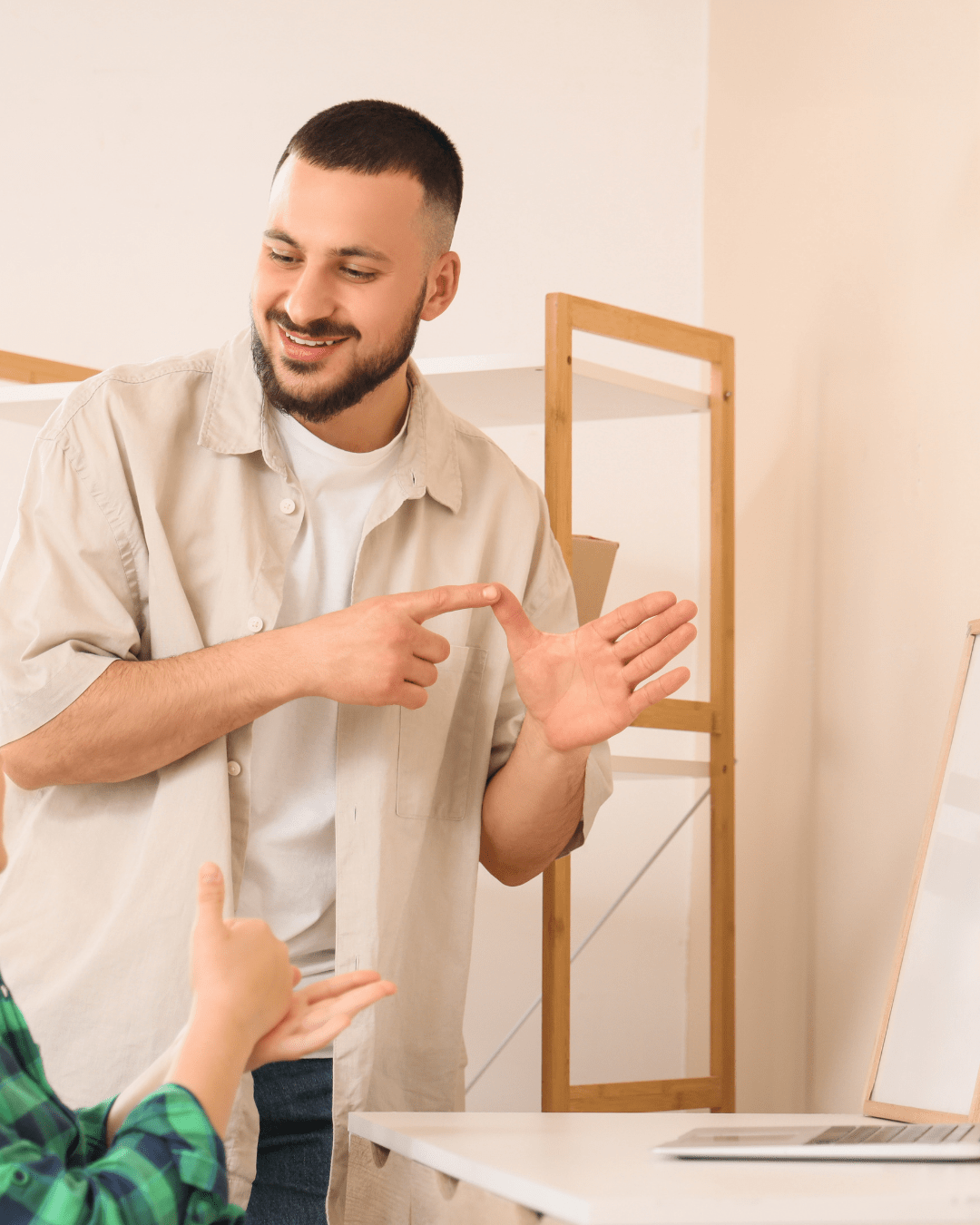 Man smiling and pointing to his hand while teaching sign language to another person indoors.