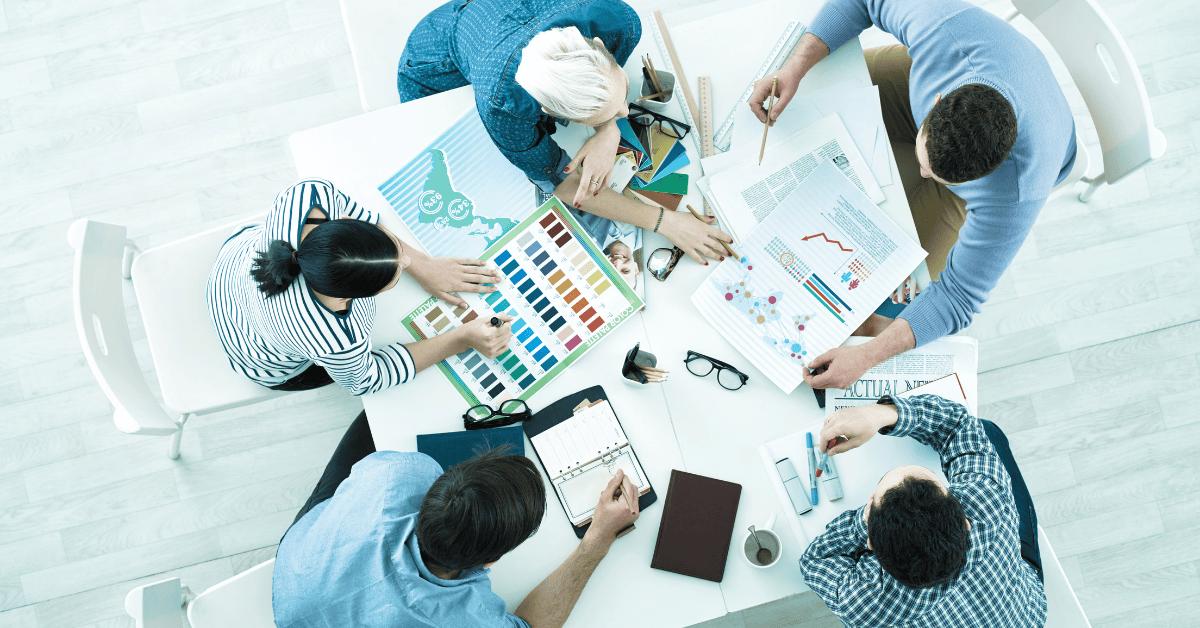 A group of six people sit around a white table engaged in a collaborative work session. They are reviewing charts, using color samples, and taking notes. Various office supplies are scattered on the table. The scene is viewed from above.