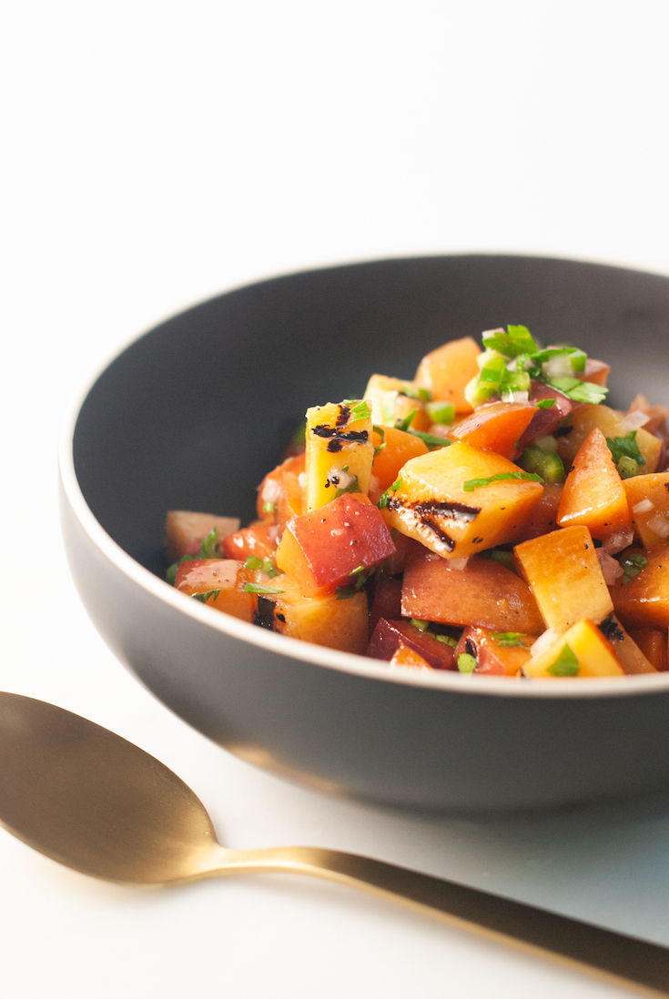 Black bowl filled with diced grilled peaches and jalapeno, with a gold spoon.