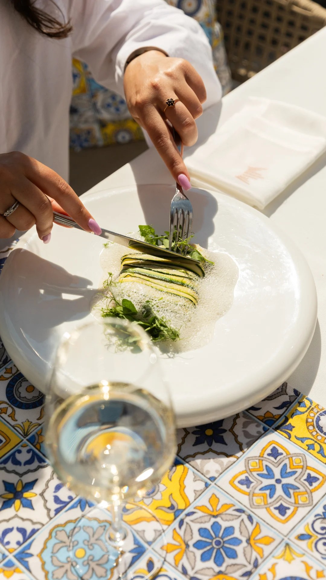 A person using a fork and knife to cut into layered zucchini on a white plate, with a glass of wine in the foreground.