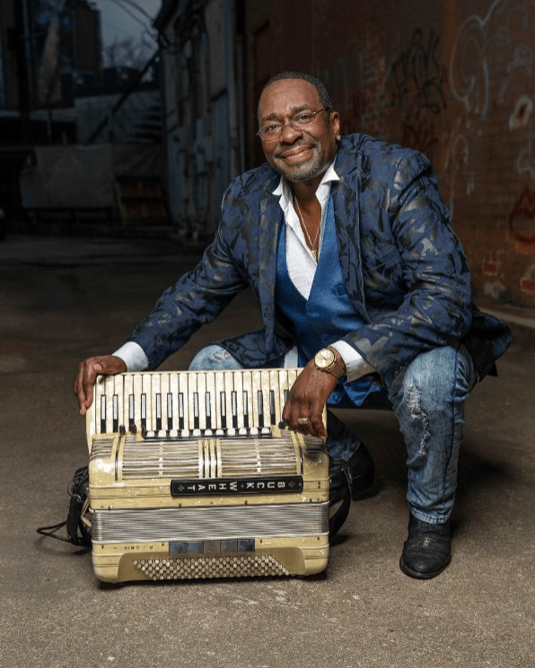 Smiling man kneeling beside a vintage accordion in an alley, wearing a blue patterned jacket