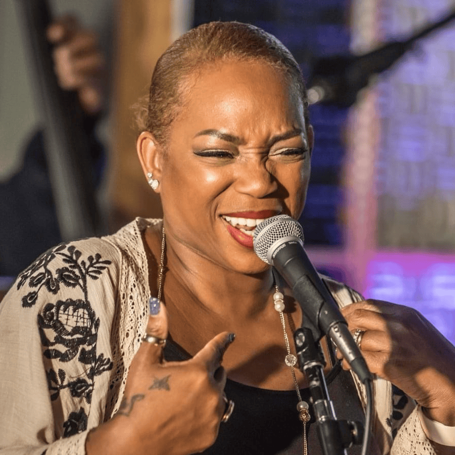 Female singer on stage, smiling and singing into a microphone, wearing a beige embroidered cardigan over a black dress with jewelry.