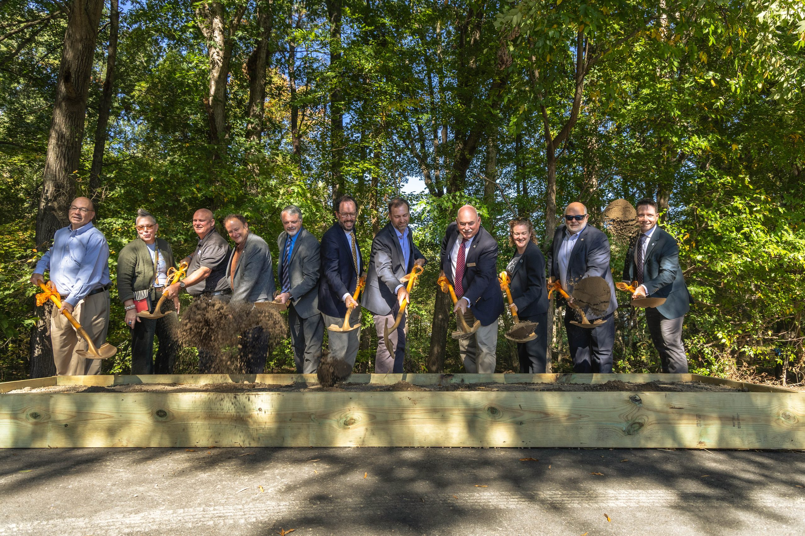 Group of suited professionals with orange-handled shovels at a woodland groundbreaking ceremony outdoors.