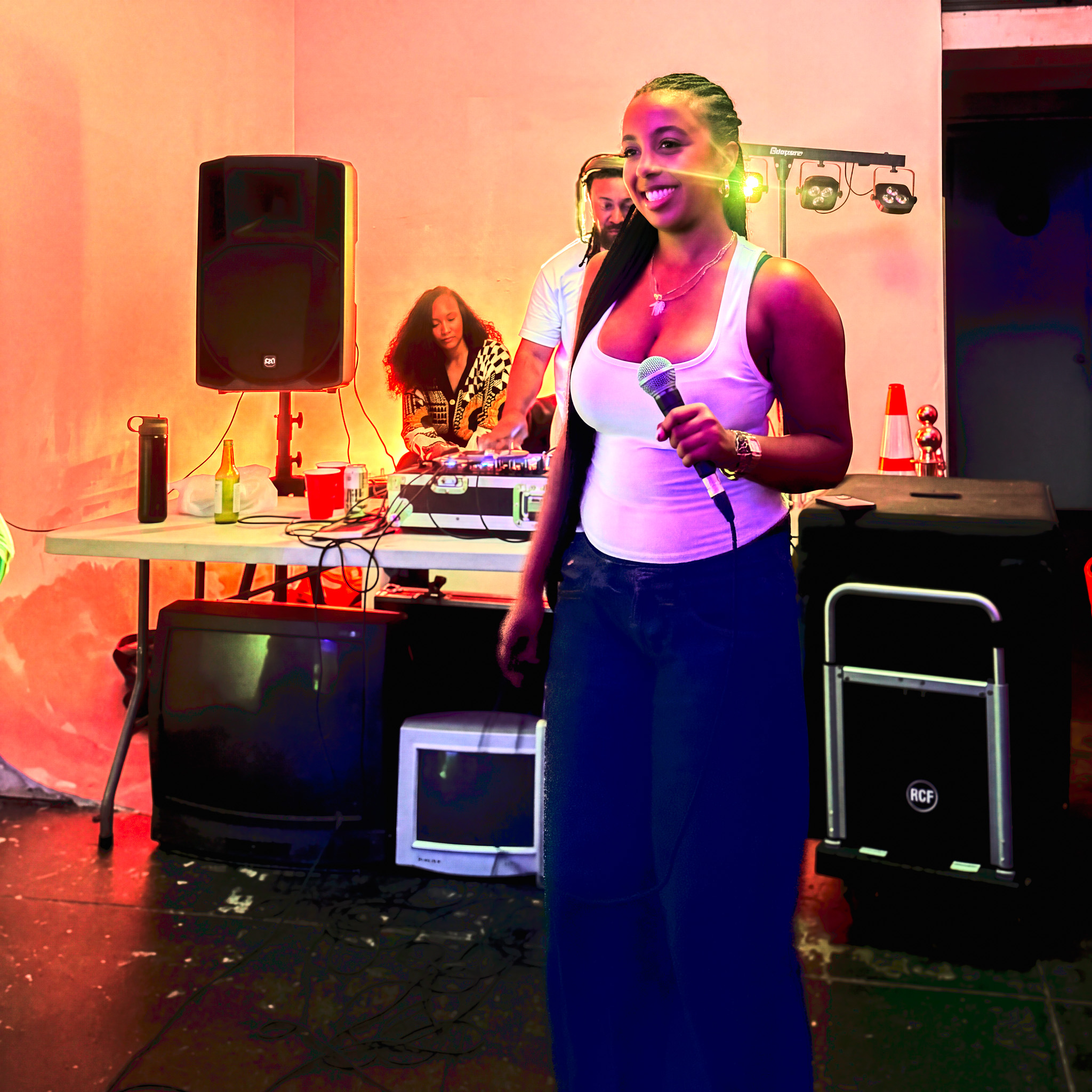 Smiling female singer in a white tank top holding a microphone on stage, with a DJ and colorful lighting in the background.