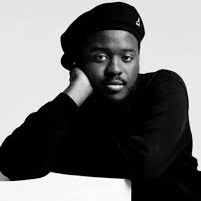 Portrait of a person wearing a beret, resting their chin on one hand, and looking at the camera in a black outfit on a light background.
