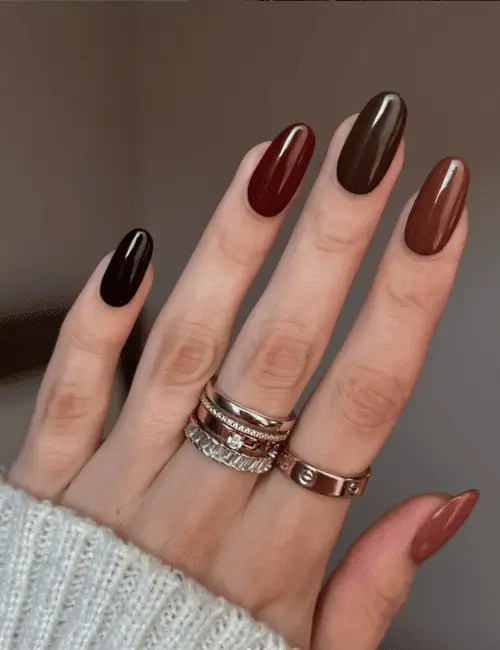 Hand with almond-shaped fall nails painted in rich earthy shades from Essie’s Boho Rodeo Collection 2025 - deep burgundy, espresso brown, terracotta, and warm nude polish, styled with gold rings and a cozy sweater.