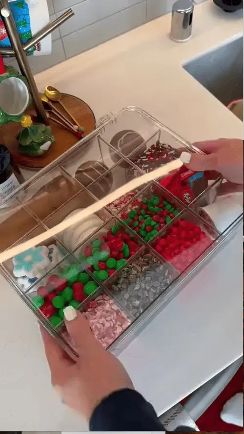 Clear acrylic organizer filled with holiday hot cocoa toppings like peppermint, marshmallows, sprinkles, and M&Ms for a neat and festive hot chocolate bar station.