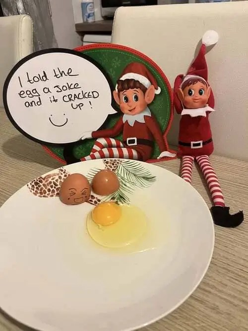 Elf on the Shelf sitting beside a cracked egg on a plate with a handwritten sign that says ‘I told the egg a joke and it cracked up’ — funny elf prank setup idea.
