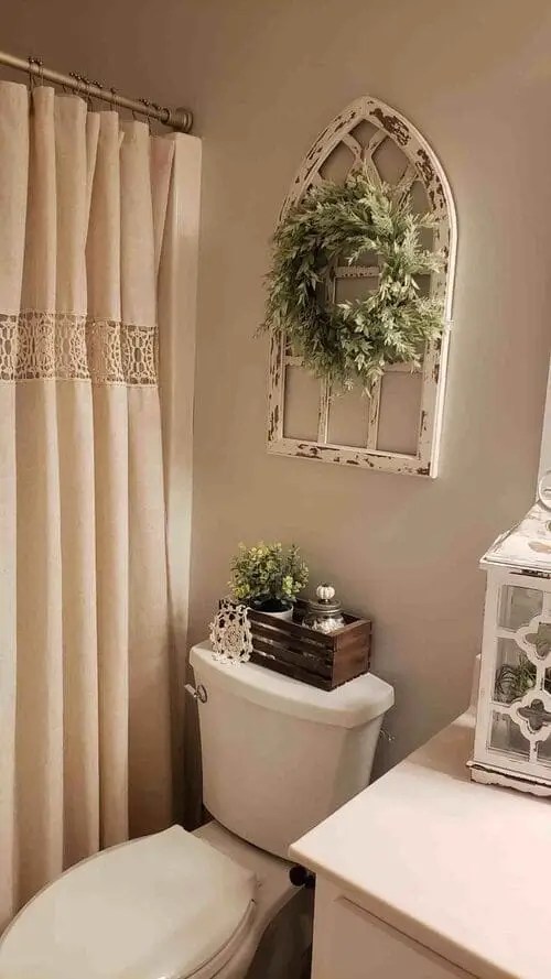 Rustic above-toilet decor featuring a vintage white window frame with wreath, wood crate with greenery and candle, and farmhouse-style bathroom accents.