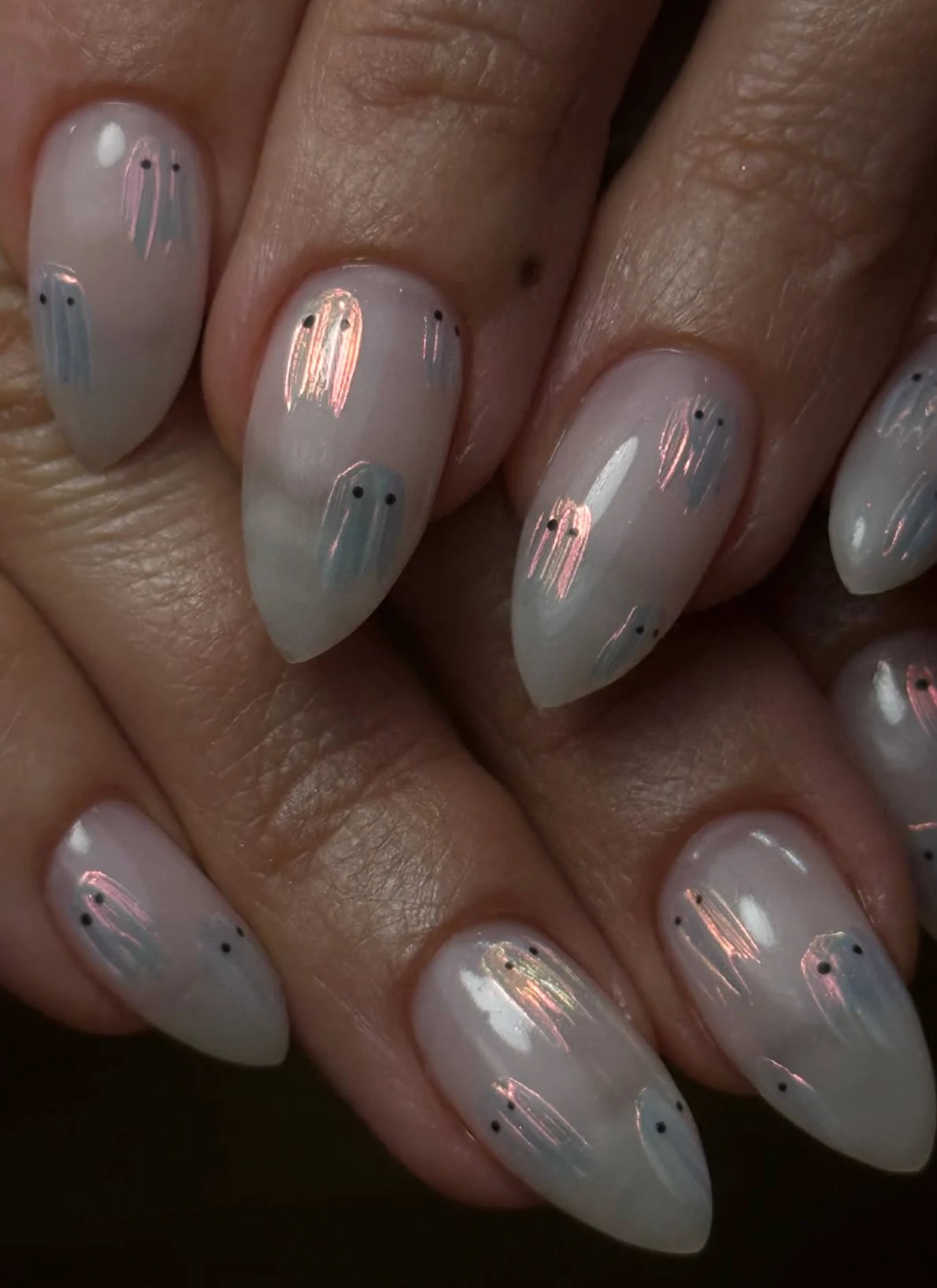 Almond-shaped milky nude Halloween nails with tiny iridescent chrome ghost designs and a glossy finish.