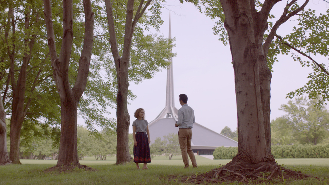 Two people stand facing a modern white church steeple among tall trees.