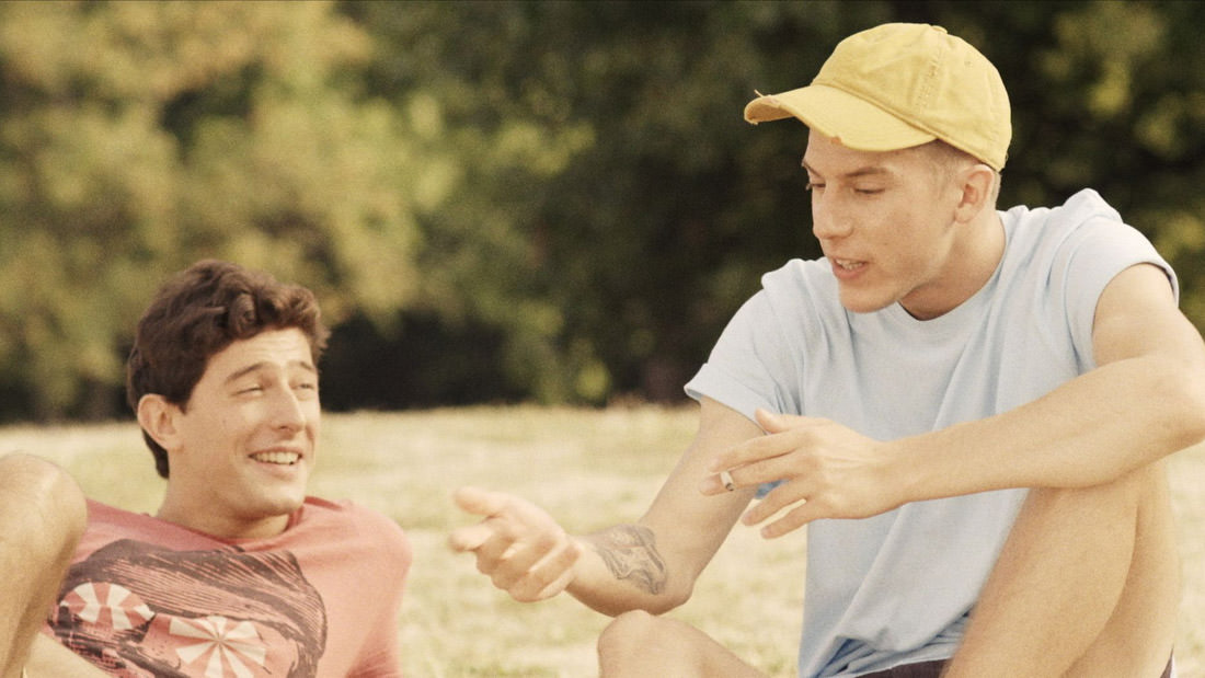 Two young men sharing a moment outdoors, one handing something to the other.