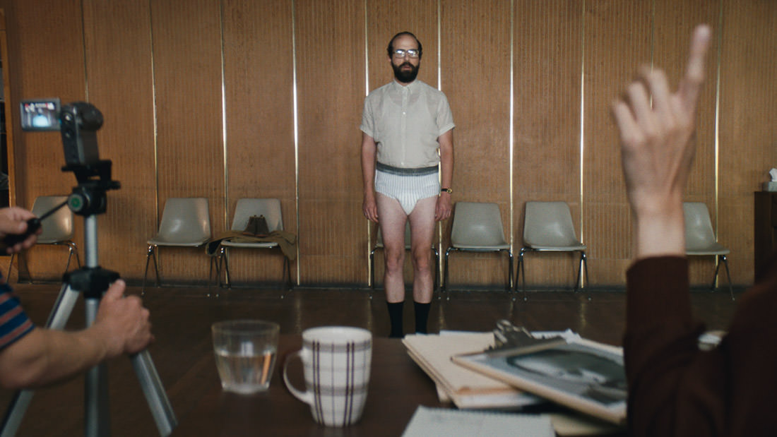 Man in a diaper standing in a room with chairs and a wooden wall.