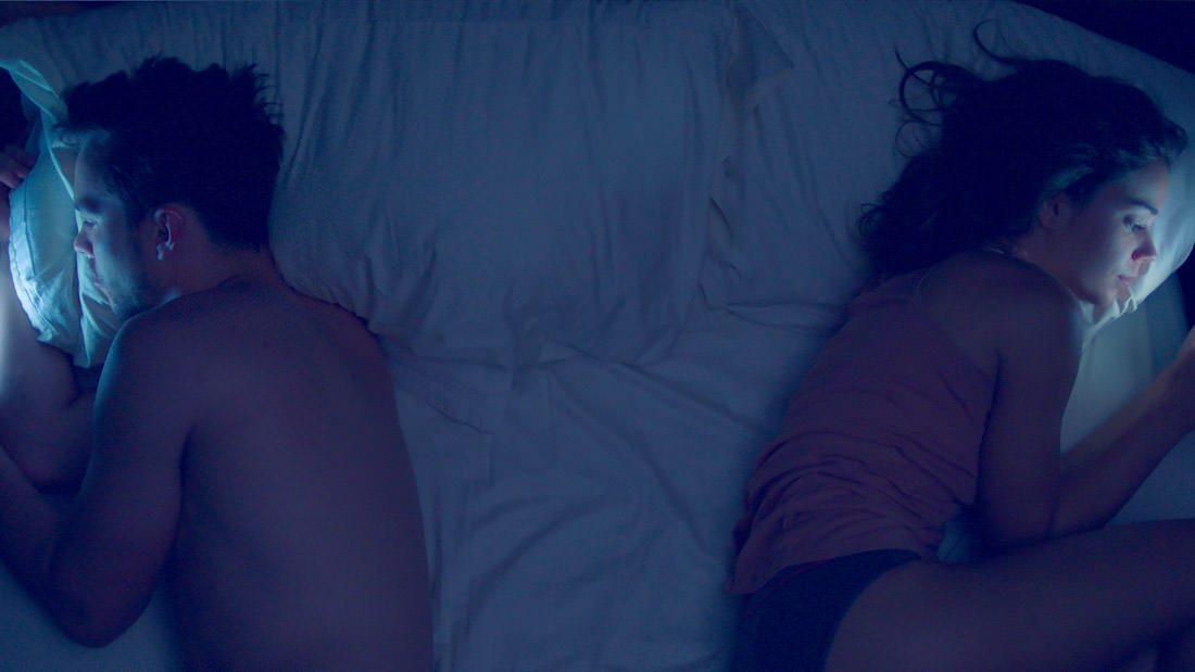 Two people lying on a bed, facing away from each other in dim lighting.