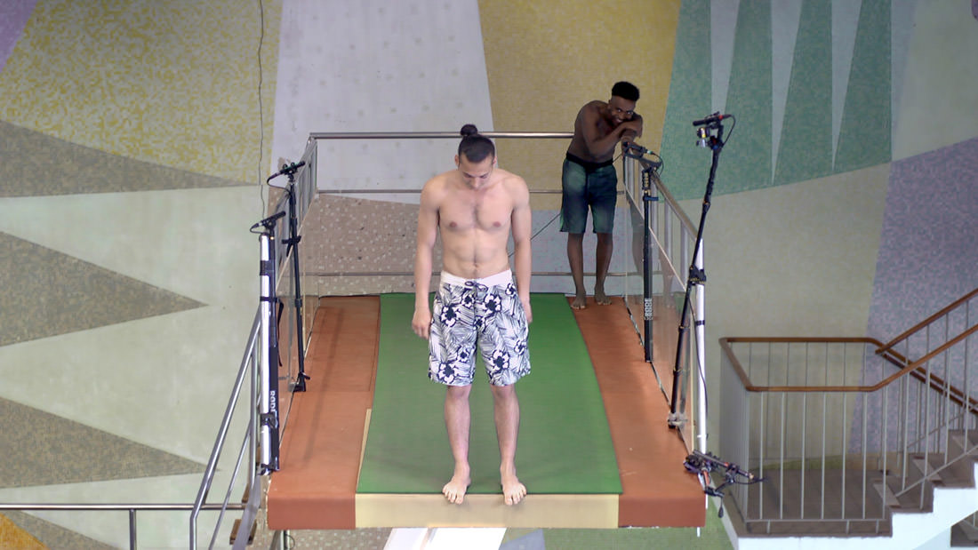 A shirtless man standing on a diving board in a swimming pool area.