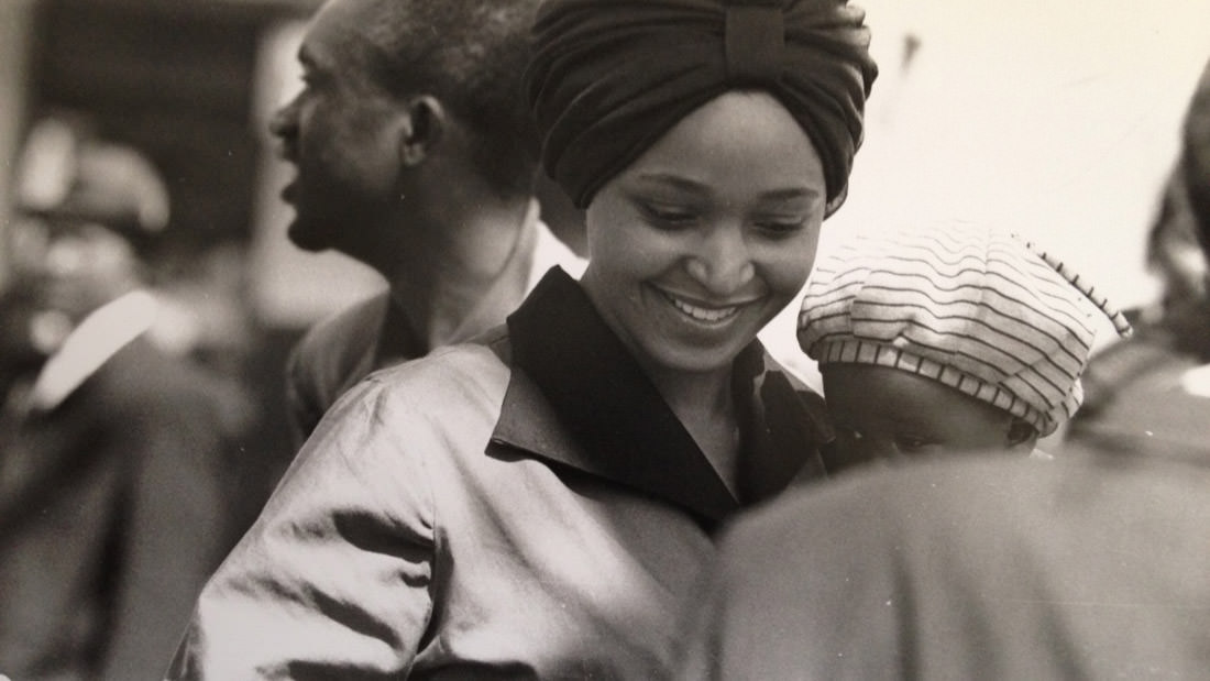 Smiling woman in a headwrap with others in background, captured in black and white.