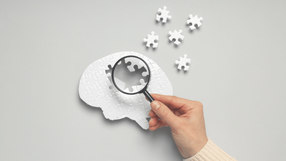 A hand holds a magnifying glass over a brain-shaped jigsaw puzzle with missing pieces, symbolising memory loss and dementia.