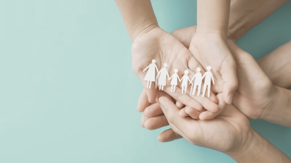 A paper cut out of a multigenerational family placed on top of six people's open hands with an aqua coloured background
