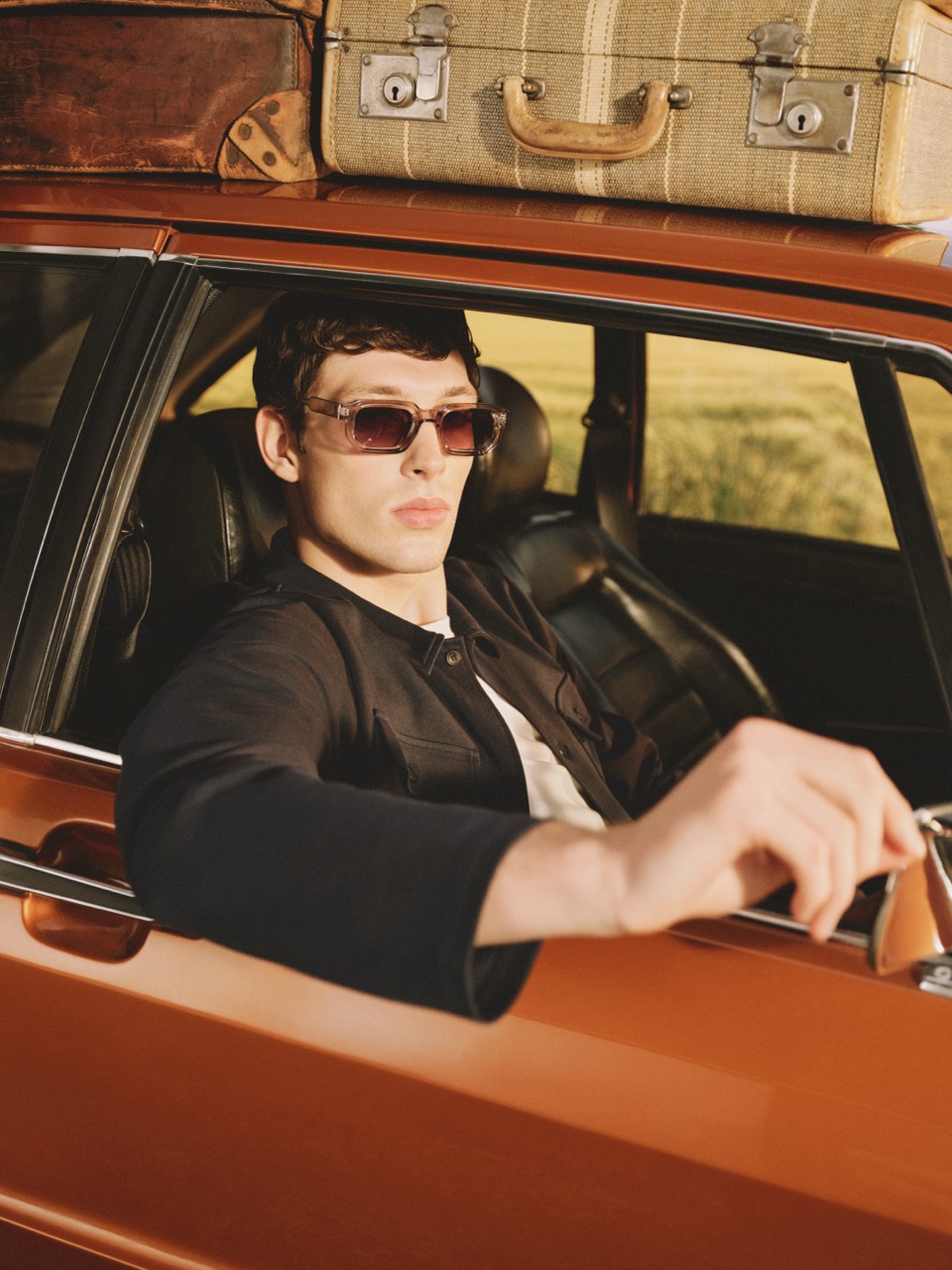 The Forge - A man wearing sunglasses sits in an orange car with his arm out of the window. Two vintage suitcases are strapped to the car roof. The background is outdoors.