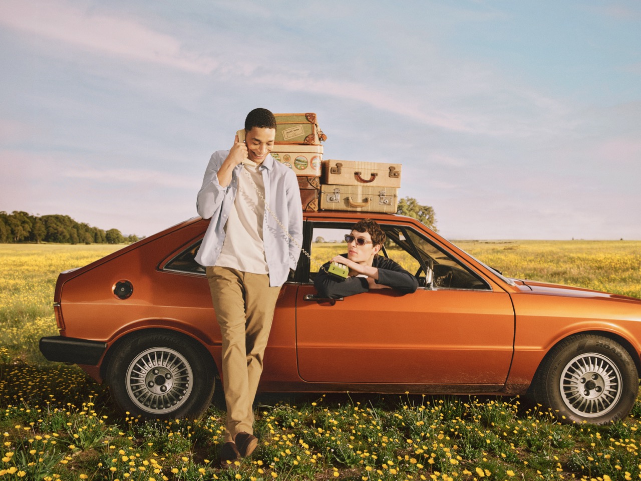 The Forge - Two people by an orange vintage car in a field of yellow flowers; suitcases are strapped to the roof. One person leans against the car talking on the phone, while the other smiles from the driver's seat.