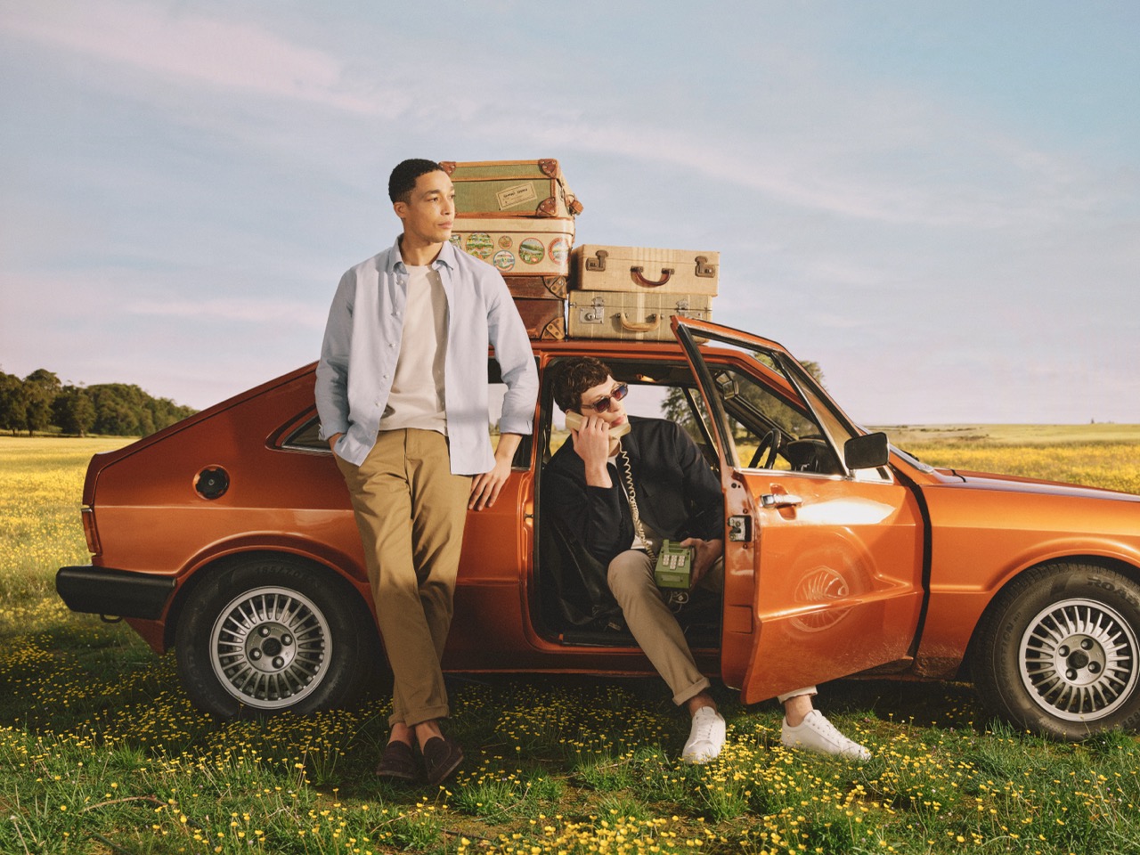 The Forge - Two men with light skin are by an orange vintage car in a field. One leans on the open door whilst the other sits inside. Several suitcases are stacked on the car roof. The field is covered with yellow flowers.