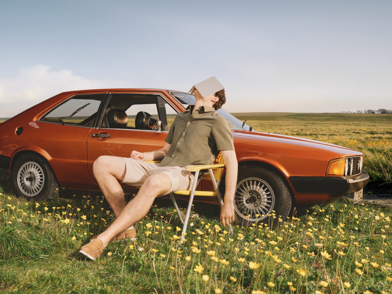 The Forge - A man in shorts and a green shirt lounges on a deckchair next to an orange vintage car in a field of yellow flowers, with a book covering his face as he relaxes.