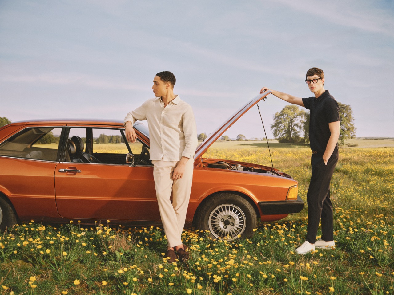 The Forge - Two men stand by an orange vintage car with its bonnet open in a field of yellow flowers. One leans on the car door, the other holds the bonnet. Trees and blue sky are visible in the background.