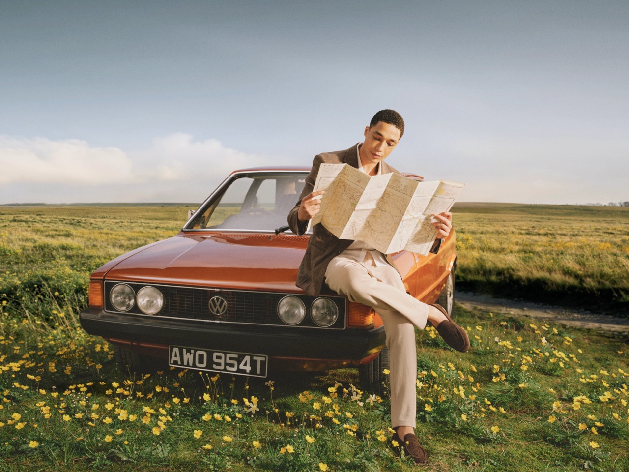 The Forge - A person sits on the bonnet of a red Volkswagen car parked in a field of yellow flowers, holding and reading a large map under a clear sky.