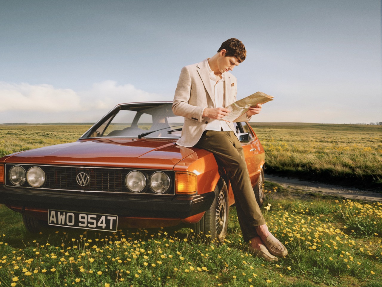 The Forge - A person in a light jacket and trousers leans on an orange classic Volkswagen car in a field of yellow flowers, reading a map under a clear sky.