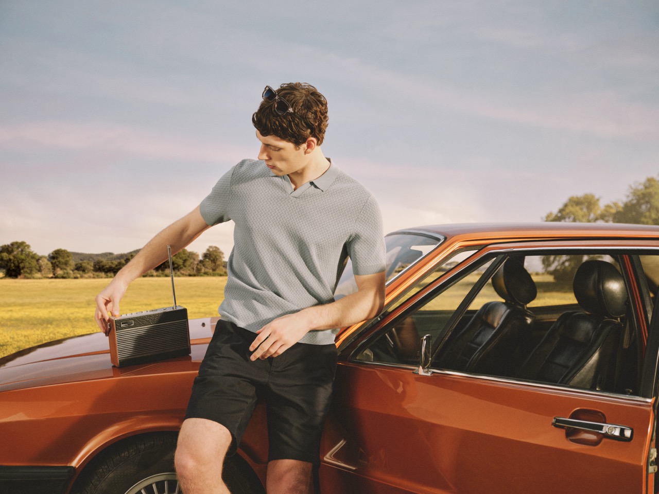 The Forge - A man in a light blue polo and black shorts leans against an orange vintage car, adjusting a radio on the bonnet. The scene is set outdoors in a field with trees under a blue sky.