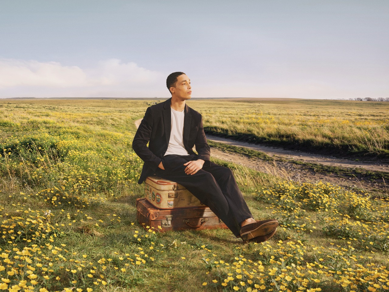 The Forge - A person in a black suit and white shirt sits on stacked suitcases in a field of yellow wildflowers, looking off into the distance under a clear sky.