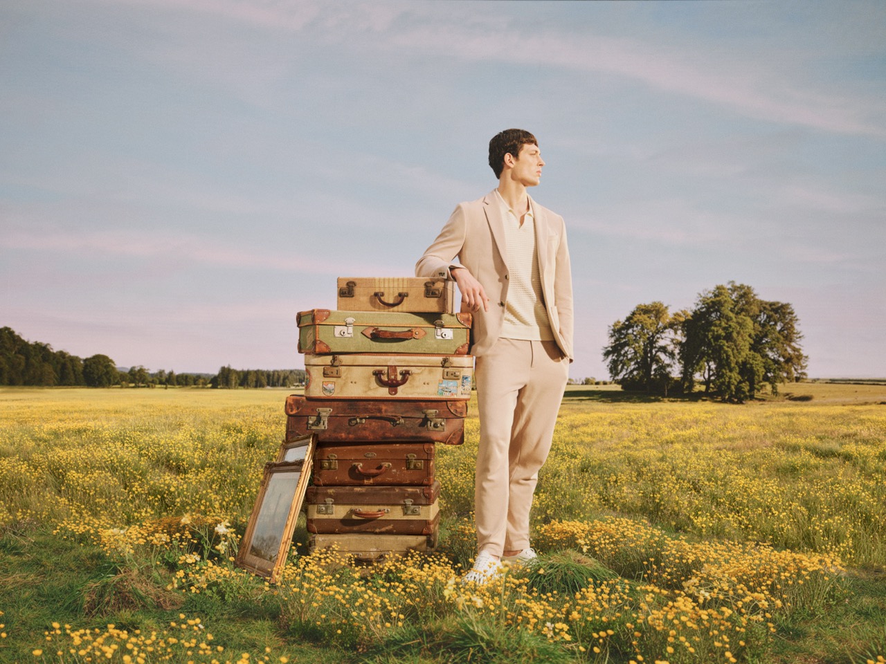 The Forge - A person in a beige suit stands in a field of yellow flowers, leaning on a tall stack of vintage suitcases, with trees and blue sky in the background.