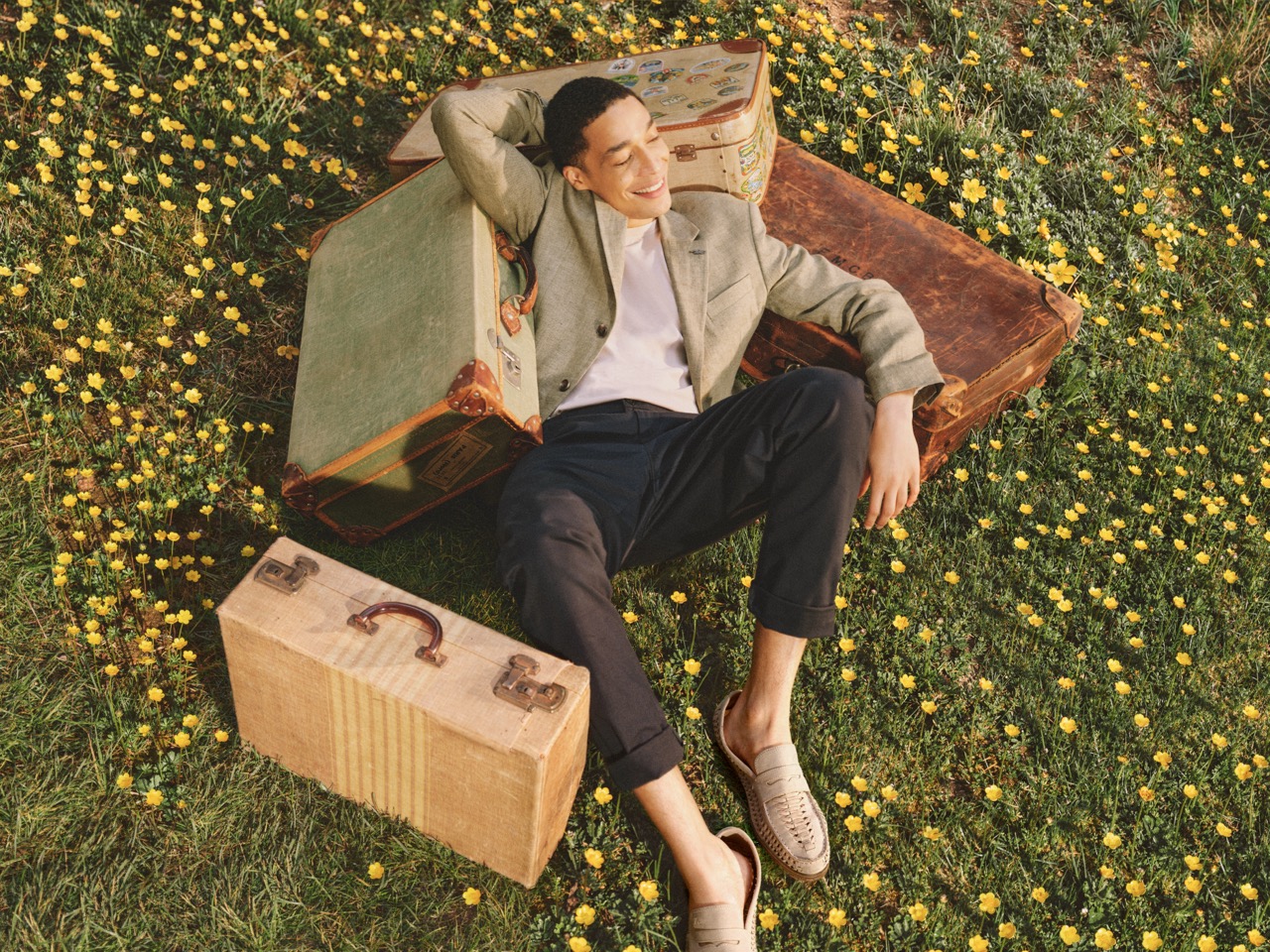 The Forge - A man relaxes on a grassy field with yellow flowers, leaning against vintage suitcases, smiling with his eyes closed.