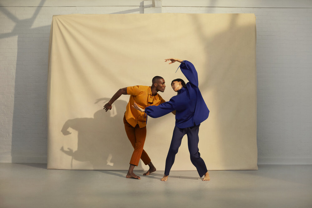 The Forge - Two dancers perform barefoot in front of a beige backdrop. One wears an orange outfit and the other a blue outfit. They lean toward each other with expressive, dynamic poses, casting shadows on the backdrop.