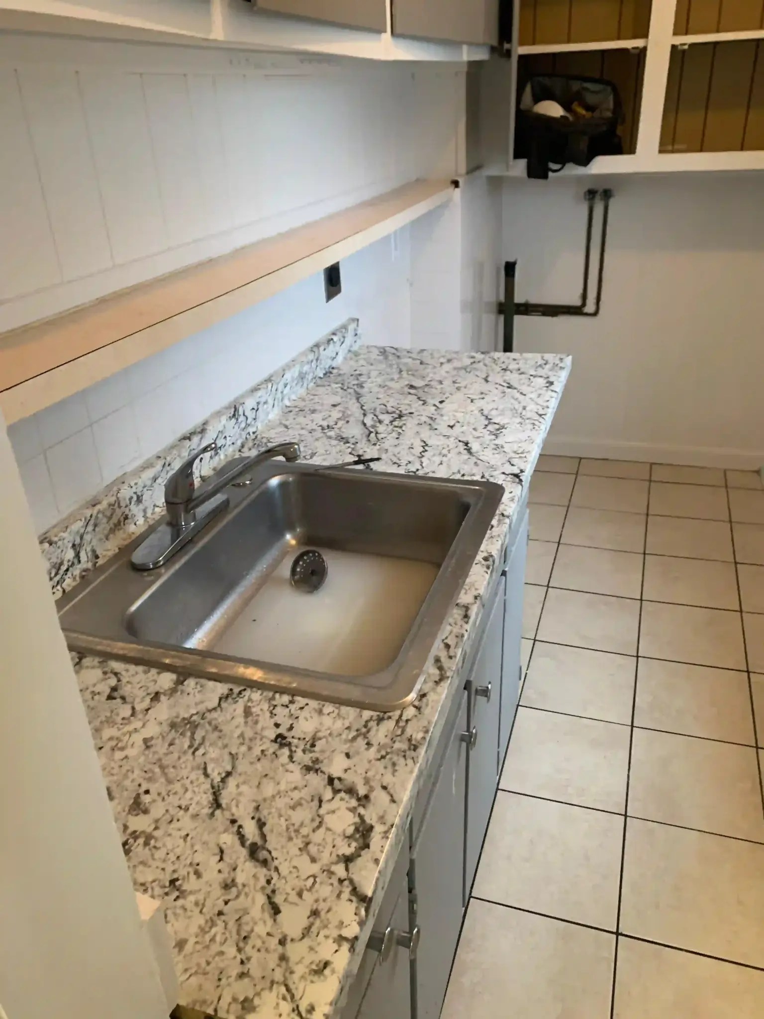 An in-progress utility room kitchen area at a New England renovation site managed by The Friendly Fixer. It features a stainless steel sink with standing water, a granite-pattern countertop, grey lower cabinets, an unfinished upper wooden shelf, and a background storage nook with exposed pipes and a tool bag.