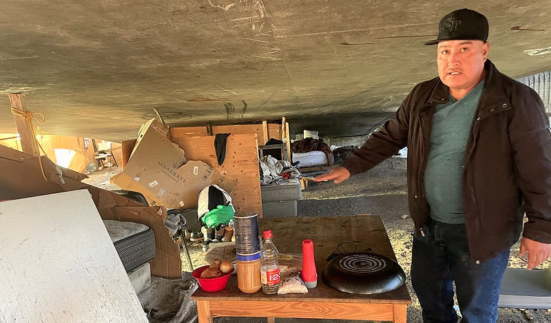A man in a black hat and jacket extends his hand to show a collection of items that make up his encampment under a San Francisco freeway.