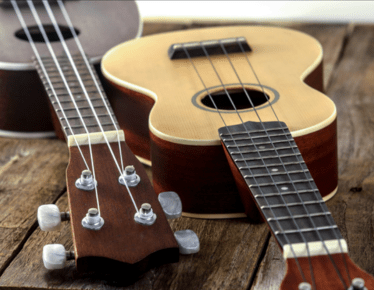 Two ukuleles resting on a wooden table, showing their necks, tuners, and strings with one light-bodied and the other darker.