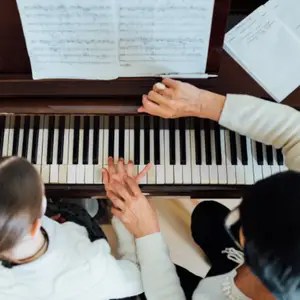 Two people at a piano; one plays while the other guides with both hands on the keys, sheet music open above the keyboard.