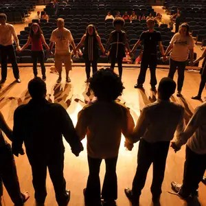 Group of people holding hands in a circle on a wooden stage in an auditorium.
