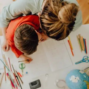 Child and adult helping with homework at a desk, surrounded by notebooks, colorful pencils, scissors, and a globe nearby.