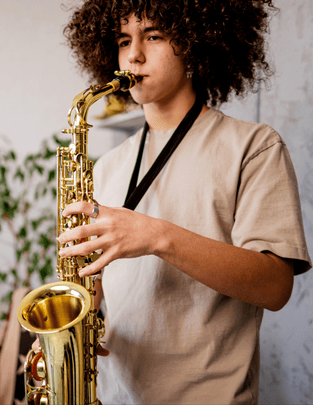 Person with curly hair playing a gold saxophone indoors, wearing a beige shirt and black strap, focusing on the instrument.