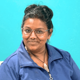Portrait of a woman with dark hair in a bun, wearing glasses and a blue jacket, standing against a bright turquoise wall.