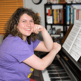 Smiling woman with curly hair sits at a piano, looking at the camera with sheet music on the stand.