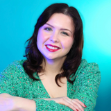Portrait of a smiling woman with dark, wavy hair in a green patterned dress against a bright blue background.
