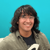 Smiling young man with wavy dark hair and glasses, wearing an olive jacket, against a teal background.