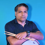 Portrait of a man with dark hair in a dark shirt, sitting against a blue wall, holding two crossed wooden drumsticks.