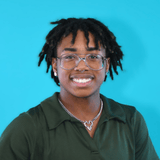 Smiling young Black man with glasses and dreadlocks, wearing a green button-down shirt against a bright blue background.